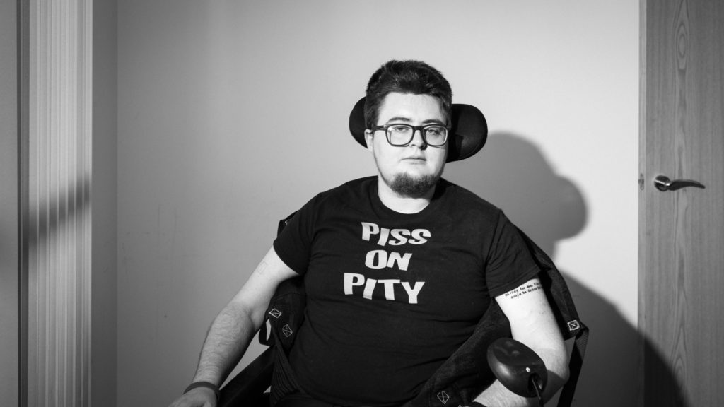 A black and white image of a young person in a wheelchair. Their head is resting on a wheelchair headreast. They have dark hair and a dark beard, and are wearing glasses. They are white, and they are wearing a black t-shirt with grey lettering, saying "PISS ON PITY"